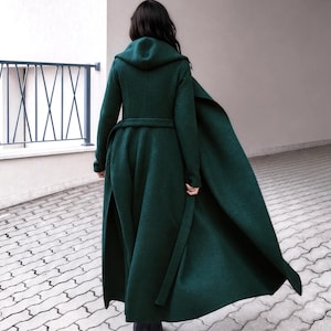 May include: A floor-length dark green coat with a hood, long sleeves, and a matching tie belt at the waist. The garment features a flared skirt design that flows outward, constructed from a textured fabric, and is worn with black boots.