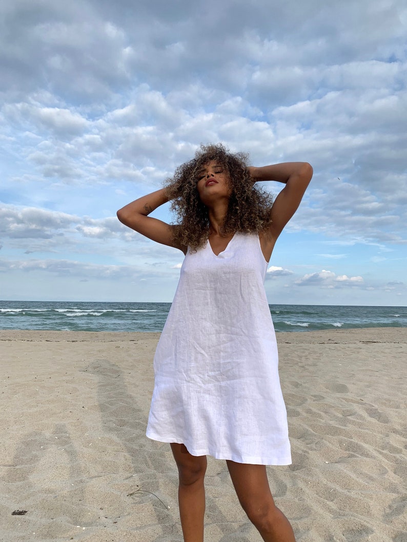 May include: A woman wearing a white linen dress with a v-neckline stands on a sandy beach with the sea in the background. The sky is cloudy.