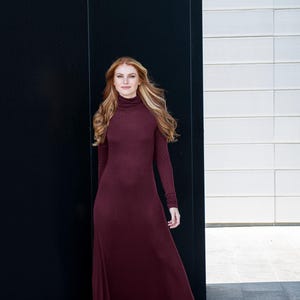 May include: A woman wearing a long, burgundy, turtleneck dress with long sleeves. She is standing in front of a black and white wall.