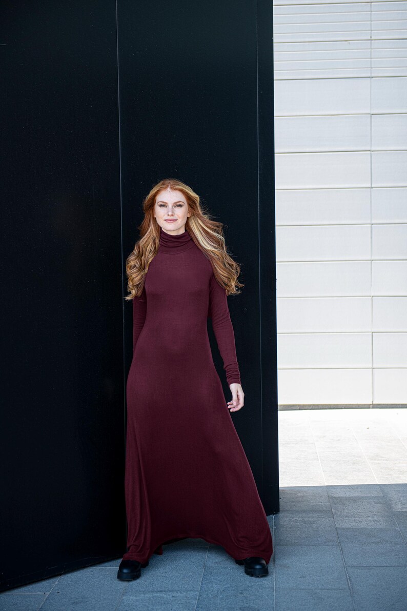 May include: A woman wearing a long, burgundy, turtleneck dress with long sleeves. She is standing in front of a black and white wall.