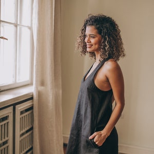 May include: A dark gray linen dress with a V-neck and pockets. The dress has a sleeveless design and a relaxed fit. The model is standing near a window with natural light. The dress is a versatile piece for casual wear.