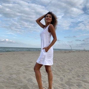 May include: A white linen dress with a sleeveless design and a pocket on the side. The dress is photographed on a woman standing on a sandy beach.