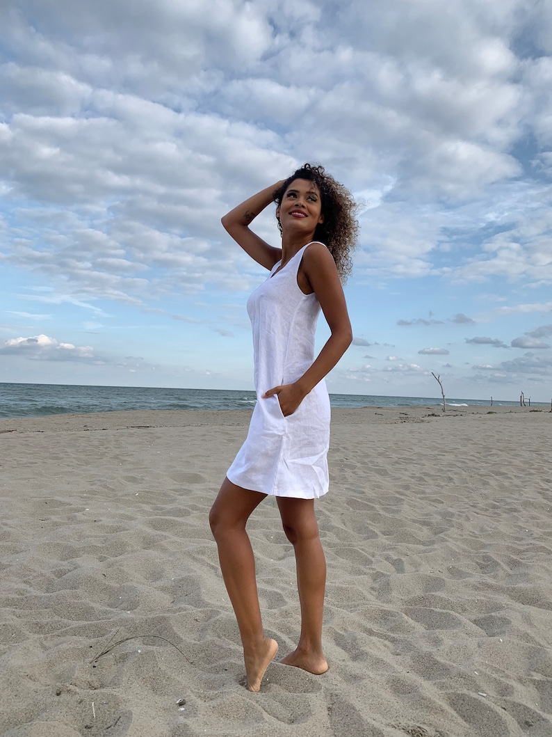 May include: A white linen dress with a sleeveless design and a pocket on the side. The dress is photographed on a woman standing on a sandy beach.