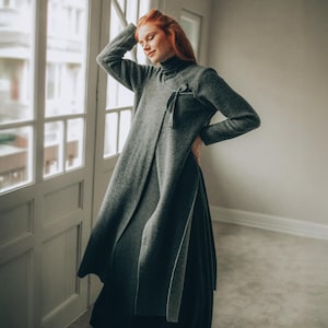 Könnte beinhalten: Ein langes, graues Rollkragenkleid mit Frontschlitz und Schleifendetail. Das Kleid hat lange Ärmel und einen fließenden Rock. Das Model steht in der Nähe eines Fensters, wobei natürliches Licht das Kleid beleuchtet.