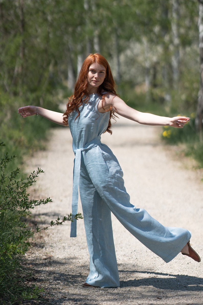 May include: A woman with long, red hair wearing a light blue linen jumpsuit with a wide leg and a belt. She is standing on a dirt path with trees in the background.
