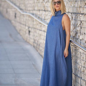 Puede incluir: Una mujer con un vestido maxi azul sin mangas, cuello alto y zapatillas blancas. Est&aacute; de pie frente a una pared de piedra.