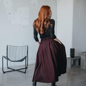 May include: A woman wearing a black long-sleeved shirt and a burgundy maxi skirt. She is standing in a room with a concrete floor and a black metal chair.