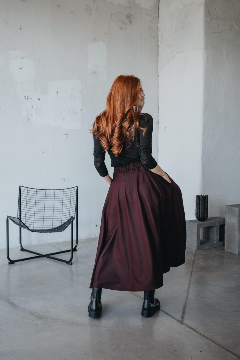 May include: A woman wearing a black long-sleeved shirt and a burgundy maxi skirt. She is standing in a room with a concrete floor and a black metal chair.