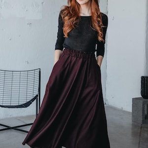 May include: A woman wearing a black long-sleeved shirt and a burgundy maxi skirt with a gathered waistband. She is standing in front of a white wall and is wearing black ankle boots.