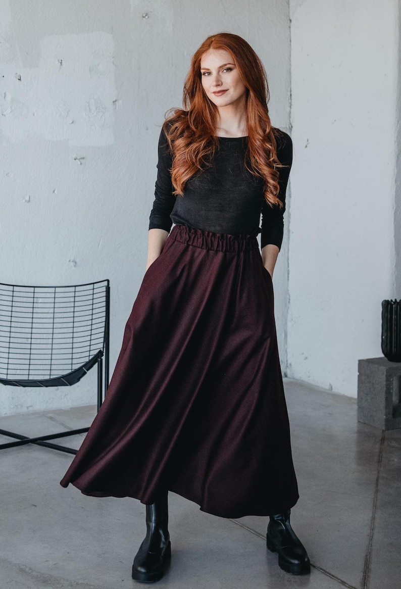 May include: A woman wearing a black long-sleeved shirt and a burgundy maxi skirt with a gathered waistband. She is standing in front of a white wall and is wearing black ankle boots.