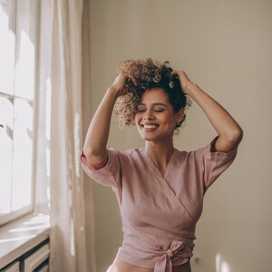 May include: A light pink wrap top with short sleeves and a tie closure, paired with white linen pants. The top is made of a soft, textured fabric. The woman is smiling and touching her curly hair.