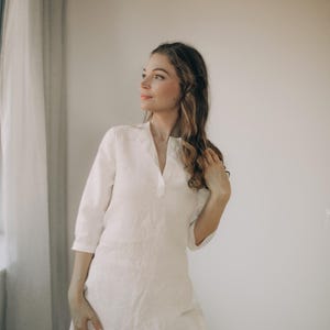 May include: A white linen tunic with a V-neck and three-quarter sleeves. The tunic is worn with light-colored pants. The model is standing near a window with sheer curtains.