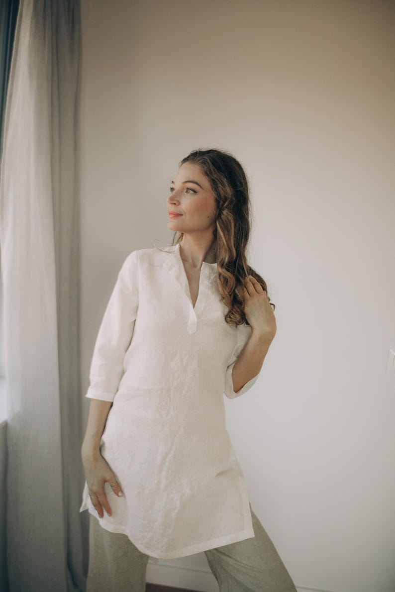 May include: A white linen tunic with a V-neck and three-quarter sleeves. The tunic is worn with light-colored pants. The model is standing near a window with sheer curtains.