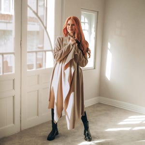 May include: A woman wearing a long, beige coat with a draped front and black boots. The coat is knee-length and has a flowing design. The background is a room with white doors and windows.