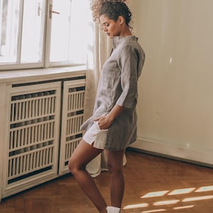 May include: A woman wearing a gray linen tunic dress with white shorts and white sneakers. The dress has a mandarin collar and three-quarter sleeves. The shorts peek out from under the dress. The woman is standing in a room with a window and wooden floor.