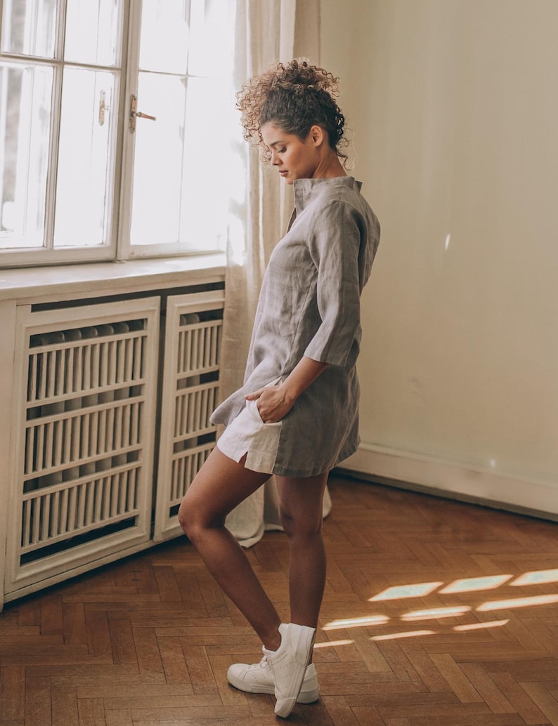 May include: A woman wearing a gray linen tunic dress with white shorts and white sneakers. The dress has a mandarin collar and three-quarter sleeves. The shorts peek out from under the dress. The woman is standing in a room with a window and wooden floor.