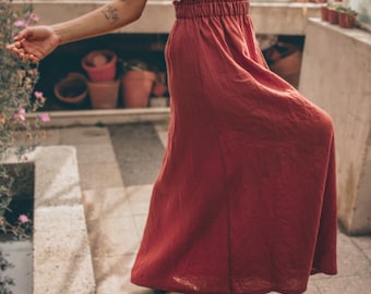May include: A sleeveless white top with a Peter Pan collar paired with a long, flowing, rust-colored skirt. The skirt has a high waist and a relaxed fit. The outfit is worn in a greenhouse setting with plants and terracotta pots in the background.