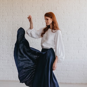 May include: A woman wearing a white blouse with long, puffy sleeves and a navy blue wrap skirt. The skirt is flowing, and the outfit is paired with white sneakers. The background is a white brick wall.