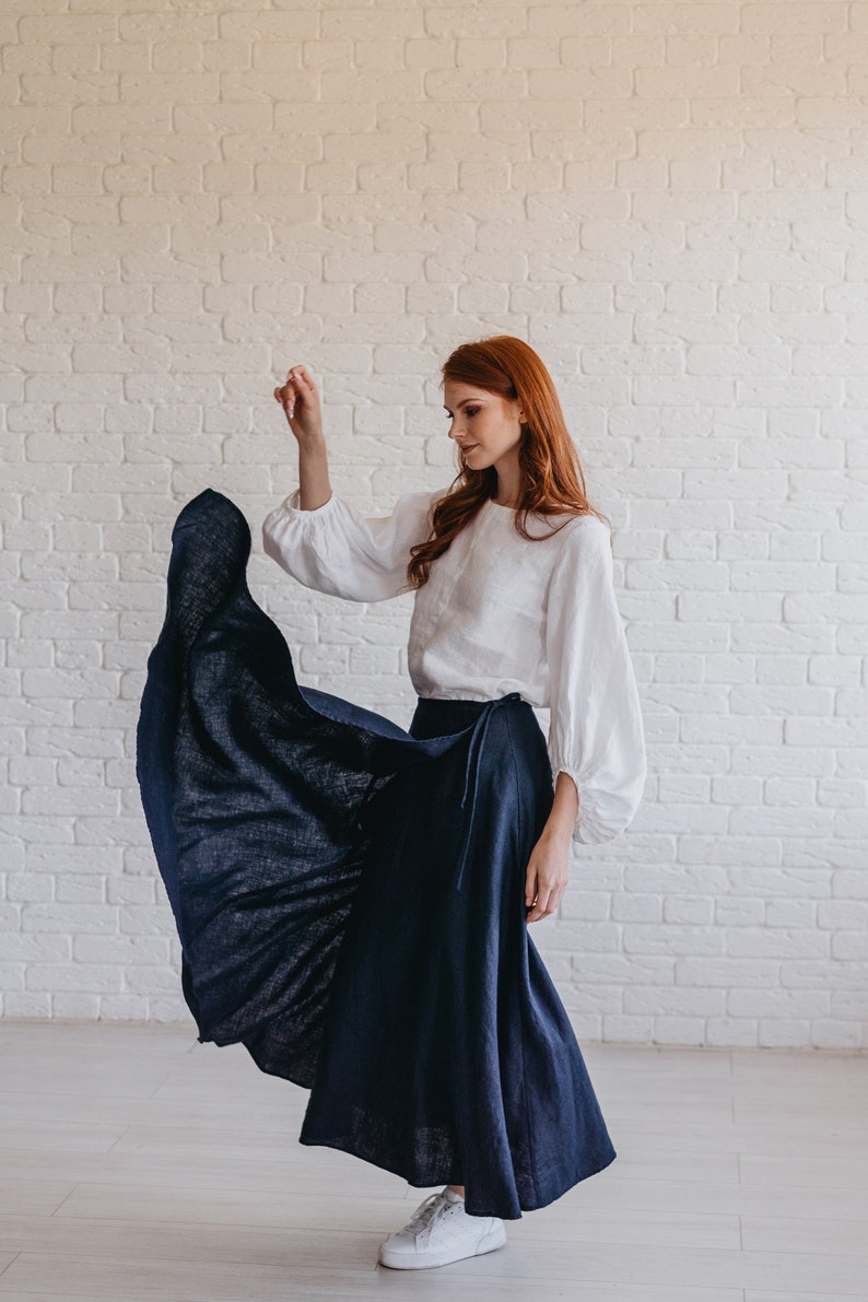 May include: A woman wearing a white blouse with long, puffy sleeves and a navy blue wrap skirt. The skirt is flowing, and the outfit is paired with white sneakers. The background is a white brick wall.