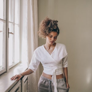 May include: A woman wearing a white wrap top with three-quarter sleeves and gray linen pants. The top ties at the waist. The pants have a drawstring. The woman is standing near a window with white curtains.
