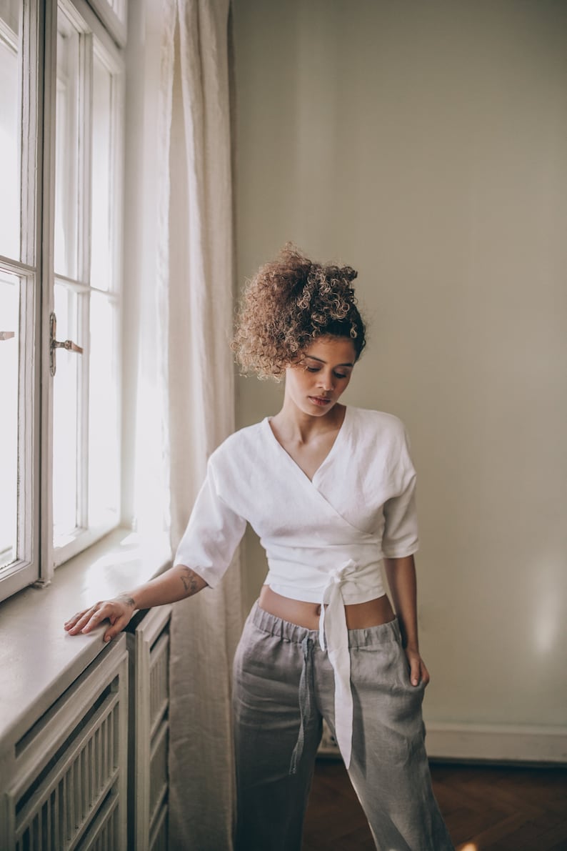 May include: A woman wearing a white wrap top with three-quarter sleeves and gray linen pants. The top ties at the waist. The pants have a drawstring. The woman is standing near a window with white curtains.