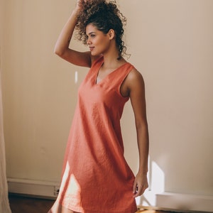 May include: A woman wearing a sleeveless, coral-coloured, linen dress with a v-neckline. She is standing in a room with a white wall and hardwood floors.