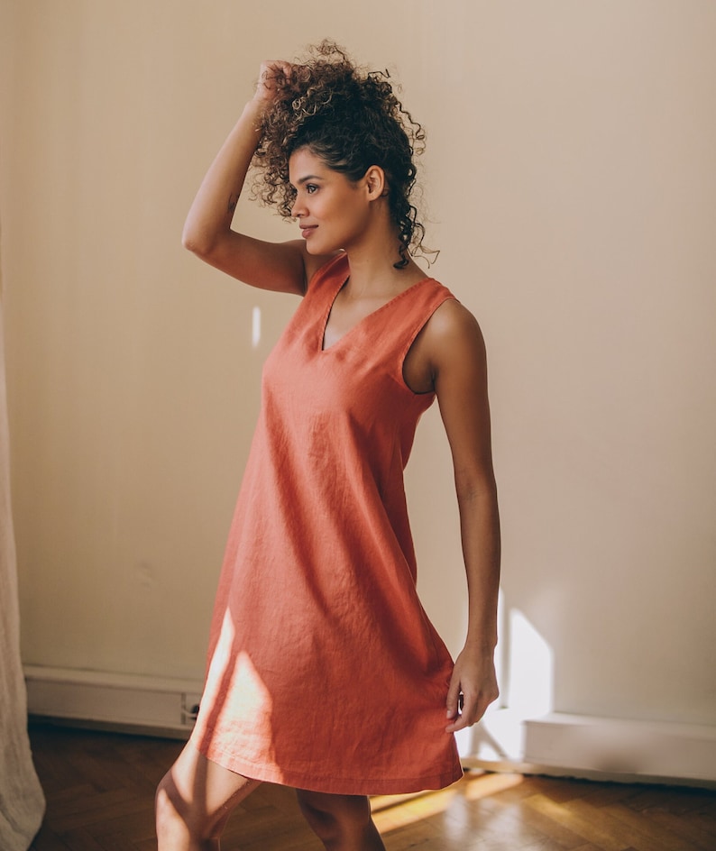May include: A woman wearing a sleeveless, coral-coloured, linen dress with a v-neckline. She is standing in a room with a white wall and hardwood floors.