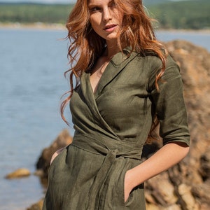 Puede incluir: Un vestido cruzado verde oliva con cintur&oacute;n anudado y mangas tres cuartos. El vestido tiene un escote en V y bolsillos. La modelo tiene el pelo largo y ondulado de color rojo. El fondo incluye una masa de agua y una costa rocosa.