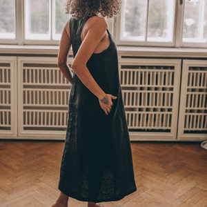 May include: A sleeveless, dark gray linen dress. The dress has a simple, flowing design and falls to the ankles. The person is standing on a wooden floor in front of a large window.
