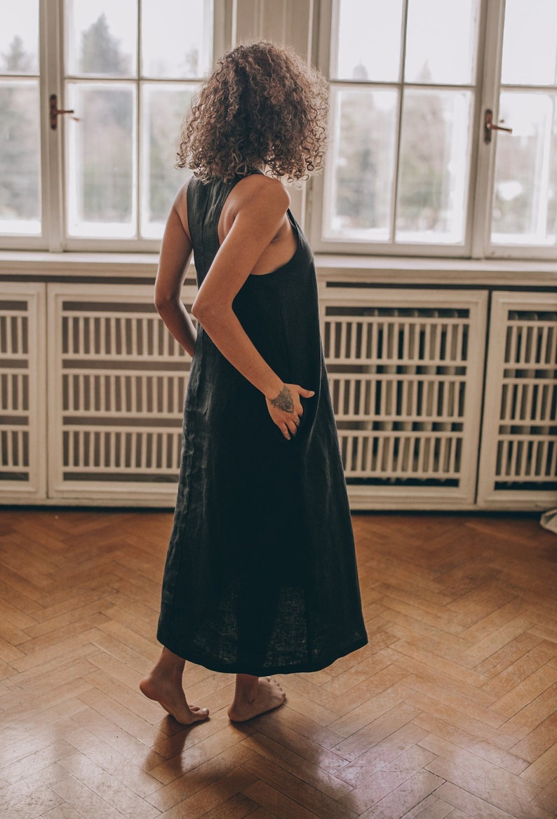 May include: A sleeveless, dark gray linen dress. The dress has a simple, flowing design and falls to the ankles. The person is standing on a wooden floor in front of a large window.