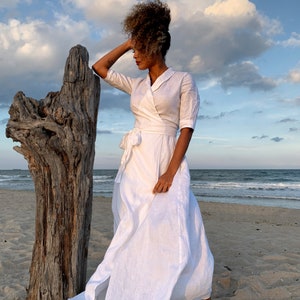 Puede incluir: Un vestido blanco cruzado con cuello y mangas tres cuartos. El vestido se ata a la cintura y tiene una falda fluida. La imagen est&aacute; tomada en una playa con un tronco de madera flotante en el fondo.