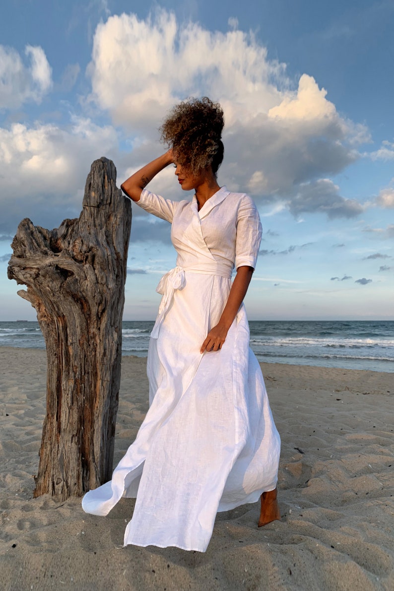 Puede incluir: Un vestido blanco cruzado con cuello y mangas tres cuartos. El vestido se ata a la cintura y tiene una falda fluida. La imagen est&aacute; tomada en una playa con un tronco de madera flotante en el fondo.