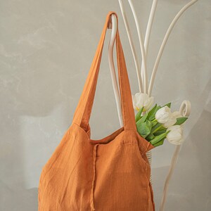 May include: A large, rust-colored tote bag made of linen fabric with a drawstring closure. The bag is hanging on a white metal stand with a bouquet of white tulips.