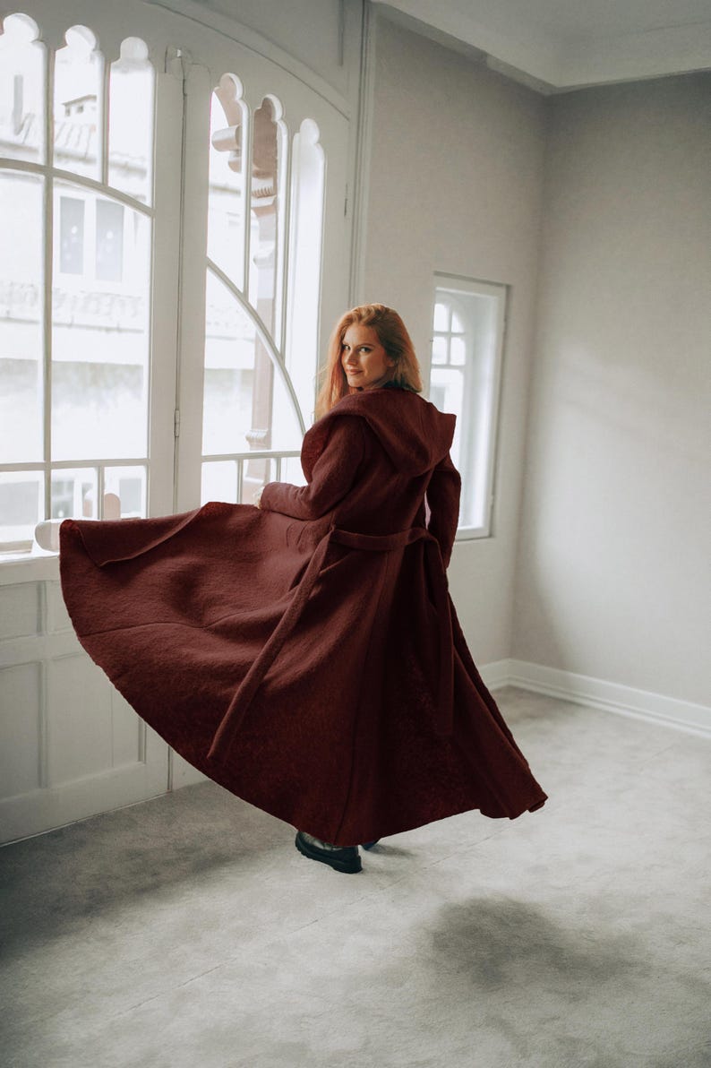 Woman wearing an elegant wool cardigan coat, standing gracefully next to a window. Handmade from a soft and cozy wool blend, this long winter coat features functional pockets, a generously sized hood, and a full-length drape for maximum comfort and style.