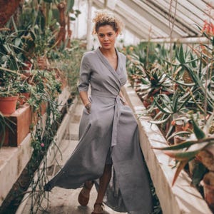 Könnte beinhalten: Ein hellgraues Wickelkleid mit passendem Gürtel und Dreiviertelärmeln. Das Kleid ist knielang und hat Taschen. Das Model trägt braune Sandalen. Der Hintergrund ist ein Gewächshaus voller Pflanzen.