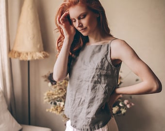 May include: A woman wearing a gray linen tank top and light pink linen pants. The top has thin straps and a square neckline. The pants have a drawstring waist. The woman has long red hair.