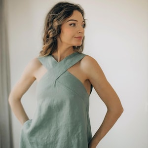 Könnte beinhalten: Ein salbeigrünes Leinenkleid mit einem einzigartigen Kreuz-Ausschnitt. Das ärmellose Kleid hat eine entspannte Passform und ein schlichtes, elegantes Design. Das Model trägt das Kleid, die Hände in den Taschen, vor einem neutralen Hintergrund.