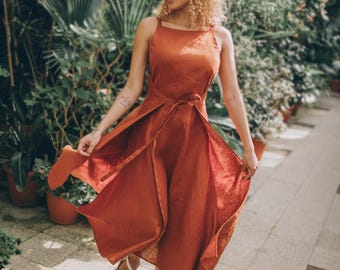 May include: A rust-colored linen dress with a tie waist and a flowing skirt. The dress has a square neckline and spaghetti straps. The person is wearing white sandals. The background is a lush garden with green foliage and potted plants.