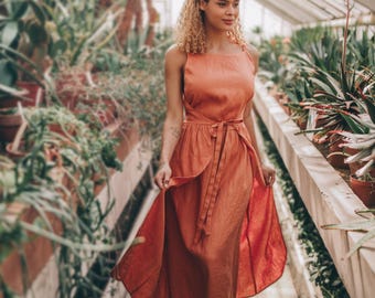 May include: A sleeveless, rust-colored dress with a tie waist and flowing skirt. The dress is worn with brown sandals. The background features a greenhouse filled with various plants and cacti, creating a natural, botanical setting.