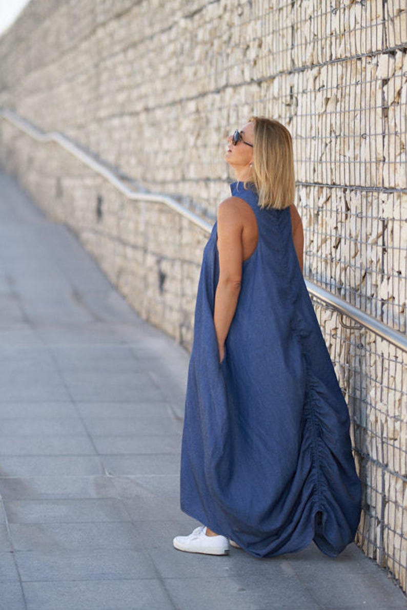 Puede incluir: Una mujer con un vestido largo de mezclilla azul con la espalda fruncida. Est&aacute; de pie en una pasarela de concreto con una pared de piedra detr&aacute;s de ella.