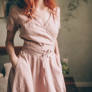 May include: A light pink linen wrap dress with short sleeves and a tie waist. The dress has a V-neckline and pockets. The fabric appears soft and flowing, suitable for warm weather. The dress is knee-length.