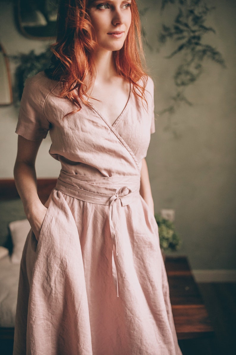 May include: A light pink linen wrap dress with short sleeves and a tie waist. The dress has a V-neckline and pockets. The fabric appears soft and flowing, suitable for warm weather. The dress is knee-length.
