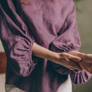 Op de afbeelding: Een pruimkleurige linnen blouse met lange, geplooide mouwen en een ronde halslijn. De blouse wordt gecombineerd met een witte broek. De stof ziet er zacht en licht uit, wat wijst op een comfortabele pasvorm.