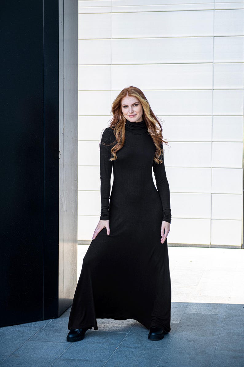 May include: A woman wearing a long, black, turtleneck dress with long sleeves. She is standing in front of a white wall and a black wall.