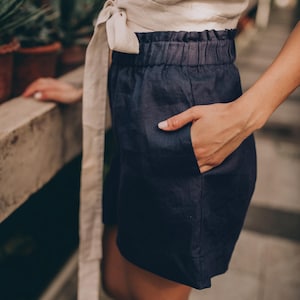 May include: Navy blue linen shorts with an elastic waistband and side pockets. The shorts are paired with a cream-colored wrap top. The outfit is casual and suitable for warm weather. The background features potted plants.