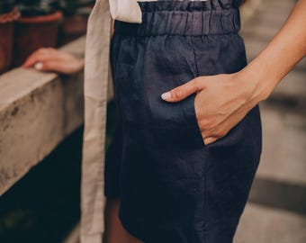 May include: Navy blue linen shorts with an elastic waistband and side pockets. The shorts are paired with a cream-colored wrap top. The outfit is casual and suitable for warm weather. The background features potted plants.