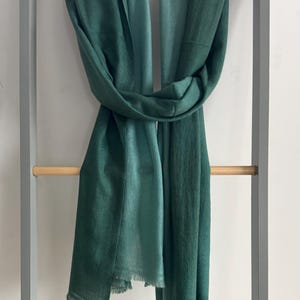May include: A green ombre scarf with a fringed edge. The scarf is folded in half and draped over a wooden rack.