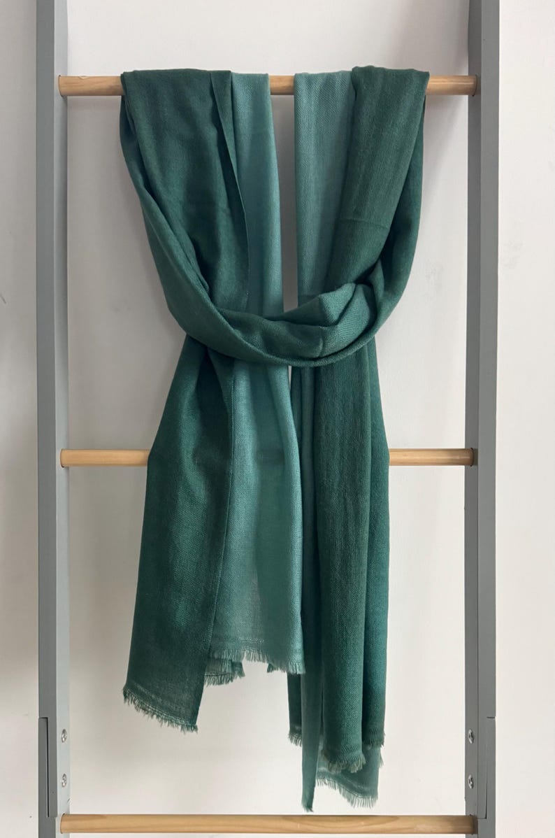 May include: A green ombre scarf with a fringed edge. The scarf is folded in half and draped over a wooden rack.