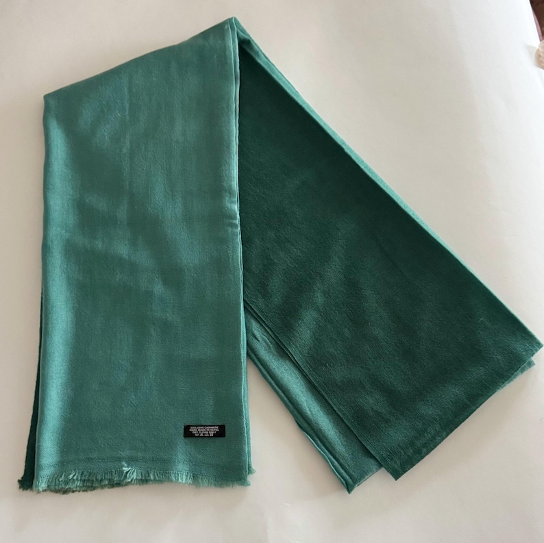 May include: A teal green pashmina scarf with a fringed edge. The scarf is folded in half and the tag is visible.