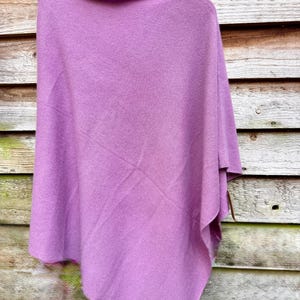May include: A solid mauve-colored knit poncho featuring a wide, draped silhouette and a soft, textured fabric surface. The garment is displayed hanging from a wooden hanger, showcasing its asymmetrical hemline and simple, clean construction.